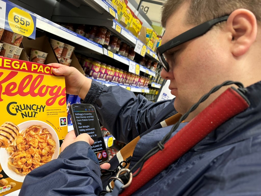 A man in a supermarket holding a stick and using his mobile phone to read the Navilens code on a packet of Crunchy Nut Cornflakes.